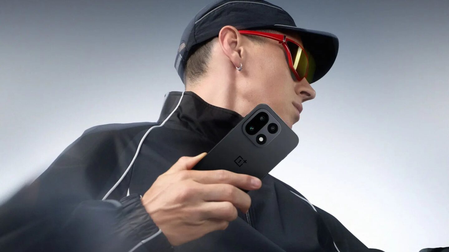 Person in a cap and sunglasses holding a black OnePlus 15